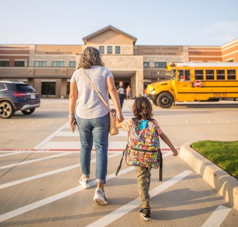 Mom walking daughter into school building with a school bus to the side. 